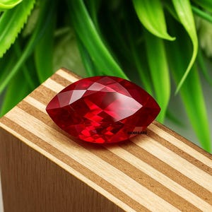 May include: A faceted, marquise-cut, deep red gemstone. The gem is displayed on a wooden block with a striped pattern. Green foliage is visible in the background. The gemstone's facets reflect light, creating a brilliant appearance.