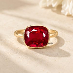 May include: A gold ring featuring a large, cushion-cut, deep red gemstone set in a smooth bezel setting. The band is polished gold, and the text GEMBRAND is printed in small black letters on the side of the setting near the base of the stone.