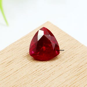 May include: A faceted, triangular-shaped, deep red gemstone. The gem is set on a light brown wooden surface. The gem has a high luster and reflects light, showcasing its many facets. The brand name "SHBRAND" is visible on the right side of the image.