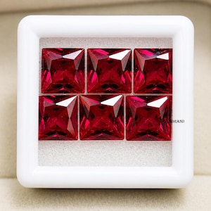 May include: Six square-cut, faceted, deep red gemstones are displayed in a white, square container. The gems are arranged in two rows of three, showcasing their brilliant cut and rich color. The gemstones are likely rubies or similar precious stones.