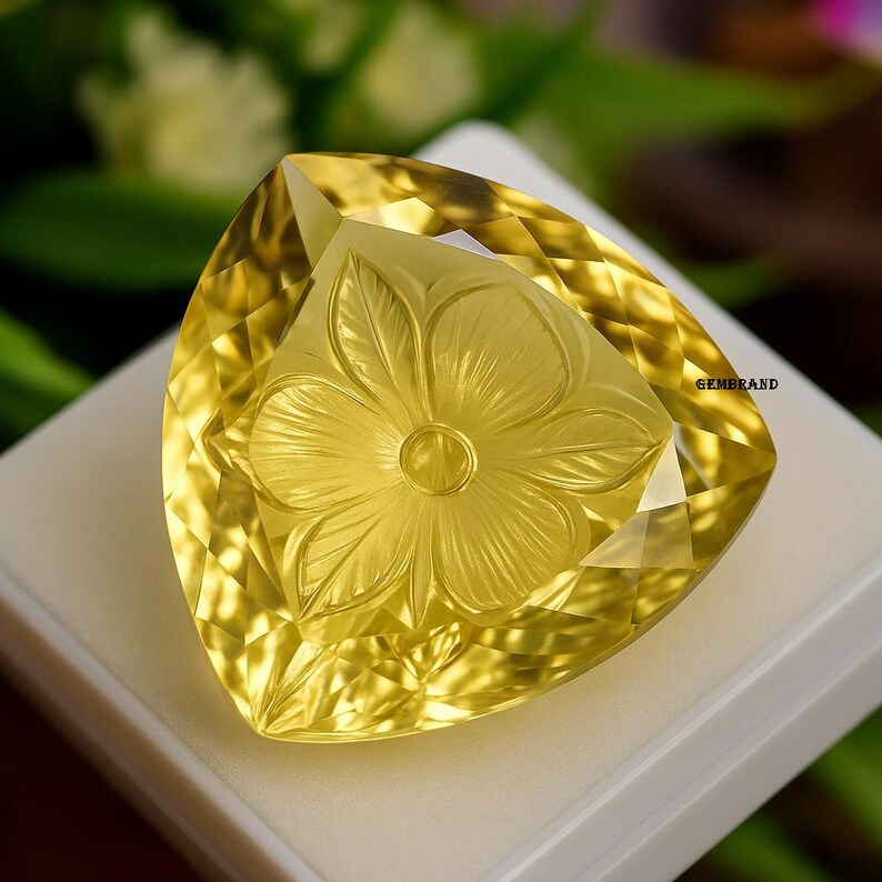 Puede incluir: Una gema amarilla triangular facetada con un dise&ntilde;o floral tallado. La gema es de color amarillo brillante y transl&uacute;cido y est&aacute; colocada sobre una superficie blanca. La palabra "GEMBRAND" es visible en el lado derecho de la gema.