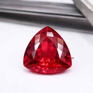 Peut inclure: Une pierre précieuse rouge foncé, taillée en forme triangulaire et à facettes. La gemme présente une taille brillante avec de multiples facettes qui réfléchissent la lumière. La pierre est posée sur un fond blanc, avec un objet métallique flou en arrière-plan.