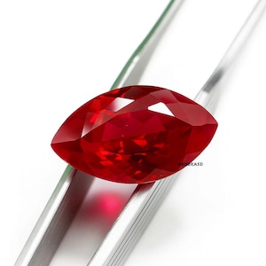 May include: A faceted, marquise-cut, red gemstone is held by a pair of tweezers. The gemstone has a high degree of clarity and brilliance, reflecting light. The tweezers are silver-colored, and the background is white.