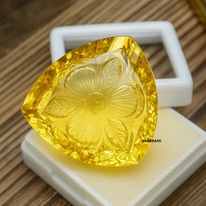 Puede incluir: Una gema amarilla triangular facetada con un dise&ntilde;o floral tallado. La gema se exhibe sobre una almohadilla de espuma blanca dentro de una caja de pl&aacute;stico blanca. La palabra "GEMBRAND" est&aacute; impresa en la caja.