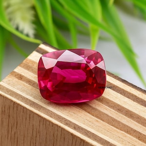 Puede incluir: Una gema de rubí facetada con corte cojín. El rubí es de un color rojo intenso y vibrante con múltiples facetas que reflejan la luz. La gema está colocada sobre una superficie de madera con follaje verde en el fondo.