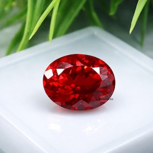 May include: A vivid red, oval-cut gemstone with multiple facets. The gem is displayed on a white surface, with the brand name "GEMBRAND" visible. The facets catch the light, creating a sparkling effect. Green foliage is visible in the background.