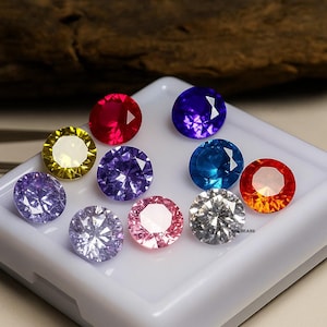 Può includere: Una collezione di dieci gemme rotonde di vari colori, tra cui rosso, viola, blu, giallo e rosa. Le gemme sono esposte su una superficie bianca, mettendo in risalto le loro sfaccettature e brillantezza. Le gemme sono probabilmente per la creazione di gioielli.