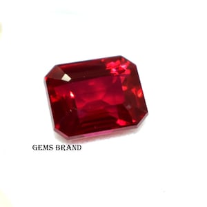Może przedstawiać: Czerwony szlachetny kamień szlifowany w kształcie szmaragdu. Kamień znajduje się na białym tle. GEMS BRAND jest nadrukowany pod kamieniem.