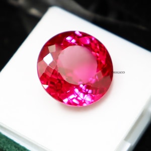 Puede incluir: Una gema rosa intenso, redonda y facetada, con talla brillante. La gema está colocada sobre un fondo blanco, con un fondo oscuro. Las facetas de la gema reflejan la luz, creando un efecto brillante. La gema es de un tono rosa vibrante.