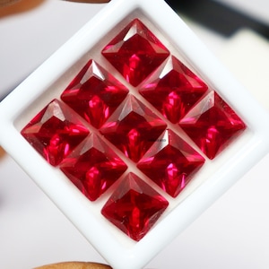 Op de afbeelding: Negen vierkante, rode edelstenen in een witte schaal. De edelstenen zijn gefacetteerd en hebben een hoge glans.