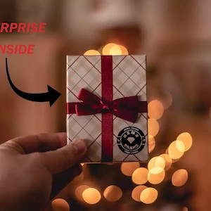 May include: A small gift box with a red ribbon and bow. The box has a white and tan plaid pattern. The words "SURPRISE INSIDE" are above the box, with an arrow pointing to it. A circular logo is on the box.