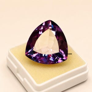 May include: A large, triangular gemstone with a faceted surface, displaying shades of purple and violet. The gem is set in a white box with a yellow interior. The gem is a striking piece of jewelry.