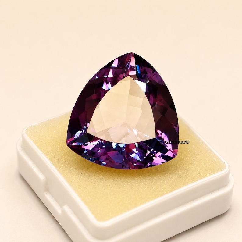 May include: A large, triangular gemstone with a faceted surface, displaying shades of purple and violet. The gem is set in a white box with a yellow interior. The gem is a striking piece of jewelry.