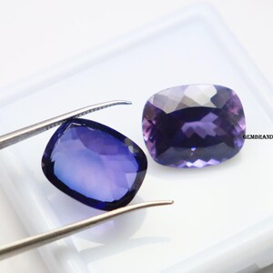 K&ouml;nnte beinhalten: Zwei violette Edelstein-Cabochons, einer mit einer Pinzette gehalten, auf wei&szlig;em Hintergrund. Die Edelsteine sind in einer rechteckigen Form mit abgerundeten Ecken geschnitten.
