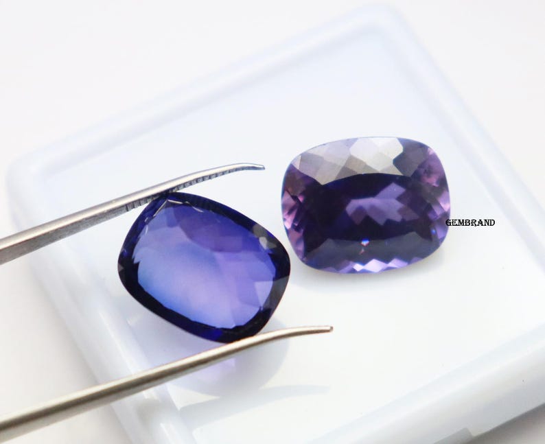 K&ouml;nnte beinhalten: Zwei violette Edelstein-Cabochons, einer mit einer Pinzette gehalten, auf wei&szlig;em Hintergrund. Die Edelsteine sind in einer rechteckigen Form mit abgerundeten Ecken geschnitten.