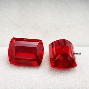 May include: Two faceted, rectangular red gemstones. One has rounded edges, the other is a half-moon shape. The gems are highly polished, reflecting light. The background is a soft, off-white colour.