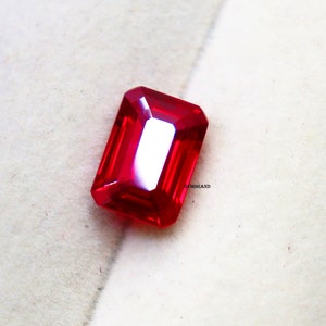 May include: A vivid, rectangular-cut, red gemstone with a polished surface. The gem has a bright, reflective quality, showcasing its facets. The gemstone is set against a neutral background, highlighting its colour and shape.