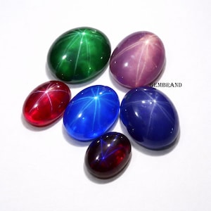 Pode incluir: Uma variedade de pedras preciosas ovais em várias cores, incluindo verde, roxo, vermelho, azul e vermelho escuro. Cada gema exibe um padrão em forma de estrela. As gemas estão dispostas sobre um fundo branco.