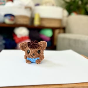 May include: A small brown crocheted plush toy with black eyes and a blue crocheted shirt. The toy is sitting on a white surface.