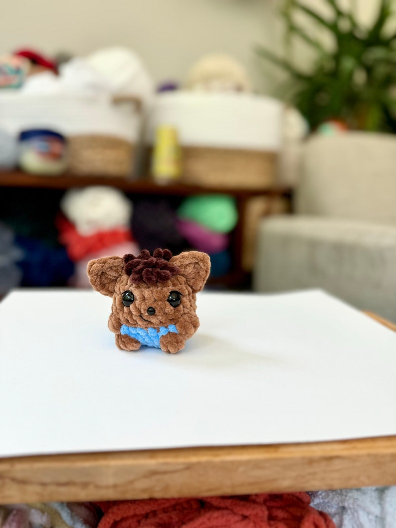 May include: A small brown crocheted plush toy with black eyes and a blue crocheted shirt. The toy is sitting on a white surface.
