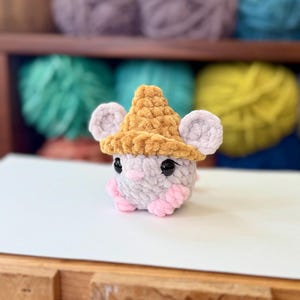 Op de afbeelding: Een kleine gehaakte muis met een bruine wollen hoed. De muis is lichtgrijs met roze accenten en zwarte ogen.