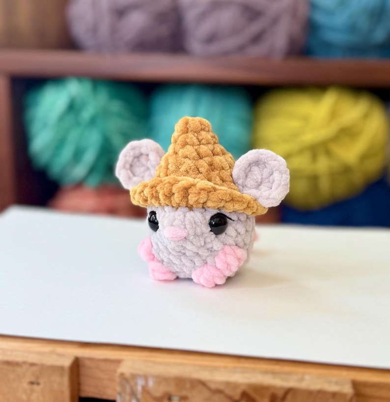 Op de afbeelding: Een kleine gehaakte muis met een bruine wollen hoed. De muis is lichtgrijs met roze accenten en zwarte ogen.