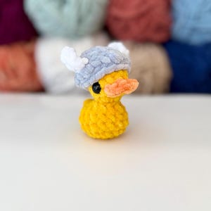 May include: A yellow plush duck with a gray and white Viking helmet and an orange beak. The duck has black eyes. The background is blurred, showing various colors of yarn.