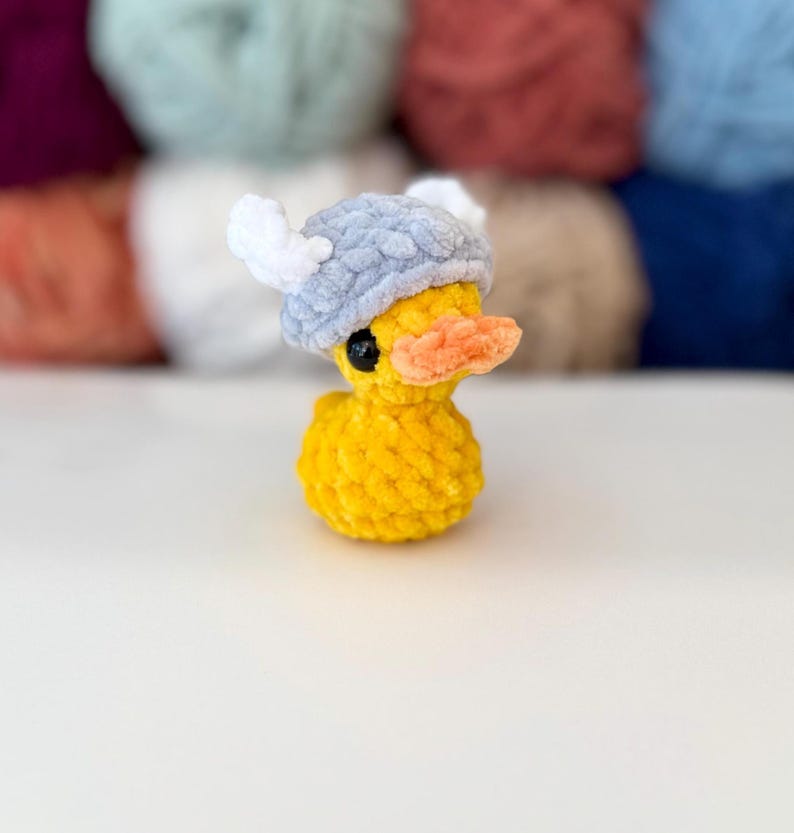 May include: A yellow plush duck with a gray and white Viking helmet and an orange beak. The duck has black eyes. The background is blurred, showing various colors of yarn.