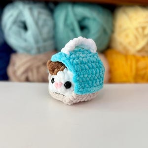 Puede incluir: Un peque&ntilde;o juguete de peluche tejido a ganchillo que representa un animal blanco y marr&oacute;n con ojos negros y nariz rosa, metido dentro de una capucha de ganchillo turquesa brillante con un borde blanco. El hilo suave y texturizado crea una forma compacta.