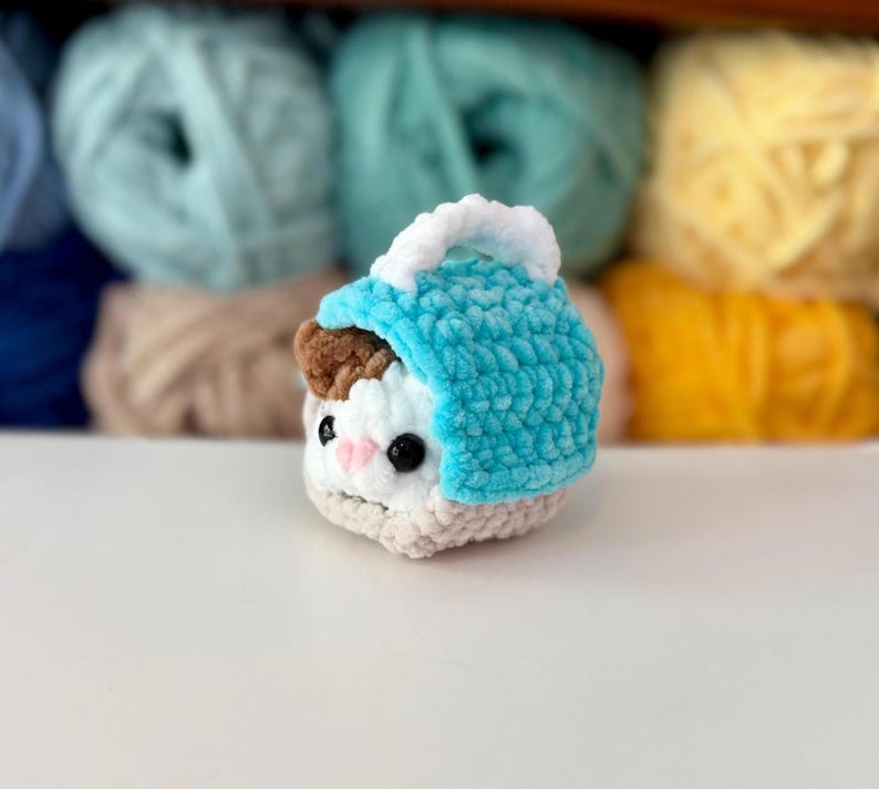Puede incluir: Un peque&ntilde;o juguete de peluche tejido a ganchillo que representa un animal blanco y marr&oacute;n con ojos negros y nariz rosa, metido dentro de una capucha de ganchillo turquesa brillante con un borde blanco. El hilo suave y texturizado crea una forma compacta.