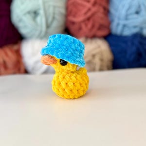 May include: A handmade yellow crochet duck wearing a blue bucket hat. The duck has an orange beak and black eyes. The background features various colours of yarn. This whimsical item is perfect for a nursery or as a unique gift.