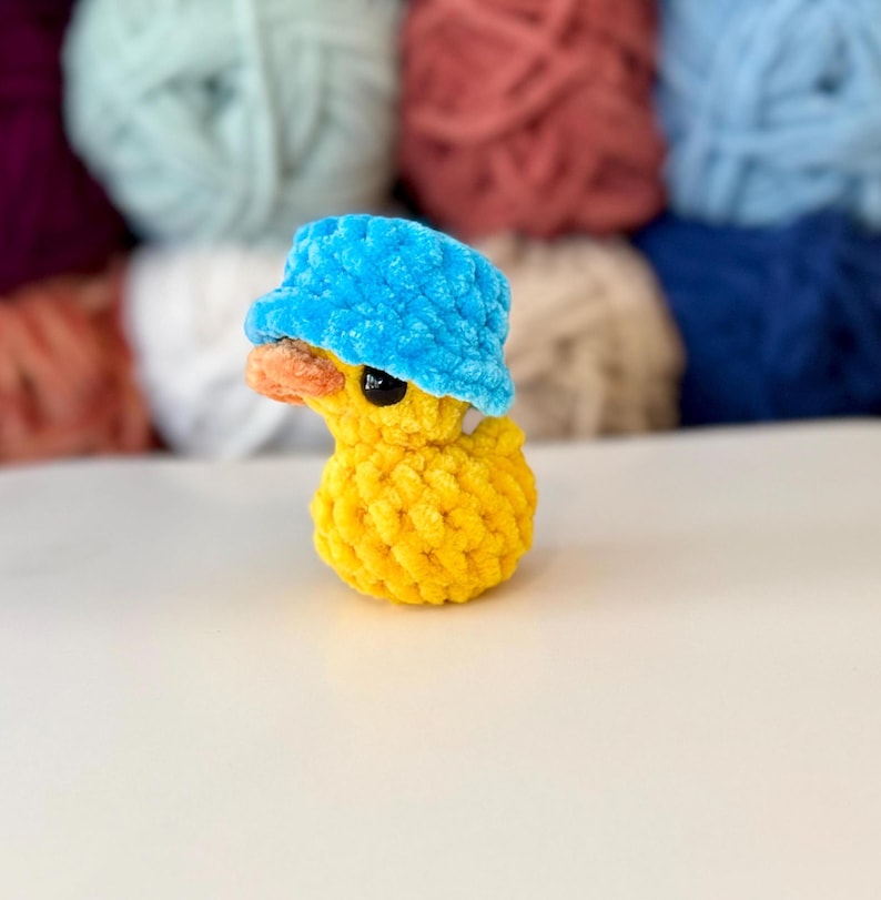 May include: A handmade yellow crochet duck wearing a blue bucket hat. The duck has an orange beak and black eyes. The background features various colours of yarn. This whimsical item is perfect for a nursery or as a unique gift.
