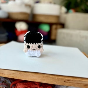 May include: A crocheted amigurumi toy of a monster with black hair, white eyes, and a pink face. The toy is sitting on a white surface.