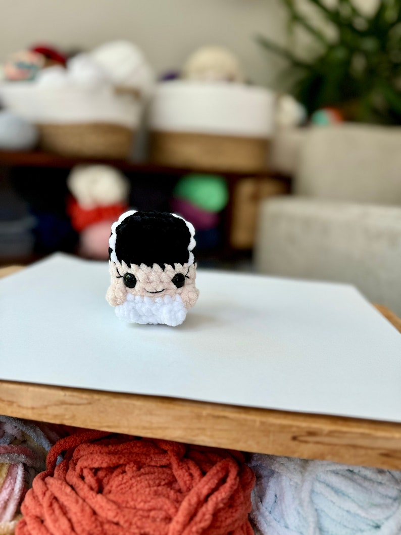 May include: A crocheted amigurumi toy of a monster with black hair, white eyes, and a pink face. The toy is sitting on a white surface.