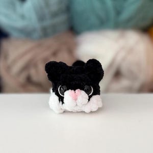 Puede incluir: Una peque&ntilde;a figura de gato tejida a ganchillo a mano, con hilo negro en la cabeza y orejas, hilo blanco en la parte inferior de la cara y las patas, y una nariz de hilo rosa claro. El gato tiene grandes ojos redondos de pl&aacute;stico negro brillante.
