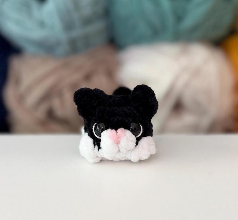 Puede incluir: Una peque&ntilde;a figura de gato tejida a ganchillo a mano, con hilo negro en la cabeza y orejas, hilo blanco en la parte inferior de la cara y las patas, y una nariz de hilo rosa claro. El gato tiene grandes ojos redondos de pl&aacute;stico negro brillante.