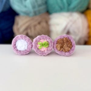 Puede incluir: Tres objetos circulares de ganchillo dispuestos en fila. Cada pieza presenta un borde exterior de color violeta claro. El objeto de la izquierda tiene un centro blanco, el del medio un centro rosa claro con un peque&ntilde;o acento verde y el de la derecha un centro marr&oacute;n.