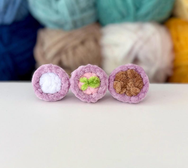 Puede incluir: Tres objetos circulares de ganchillo dispuestos en fila. Cada pieza presenta un borde exterior de color violeta claro. El objeto de la izquierda tiene un centro blanco, el del medio un centro rosa claro con un peque&ntilde;o acento verde y el de la derecha un centro marr&oacute;n.