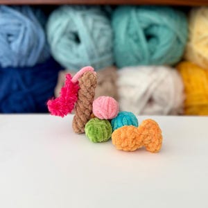 Puede incluir: Una colecci&oacute;n de peque&ntilde;as formas de peluche tejidas a ganchillo con hilo suave y texturizado. El conjunto incluye un pilar vertical marr&oacute;n con flecos rosa brillante, una esfera rosa, una esfera verde, una esfera azul y una pieza naranja con forma de pez.