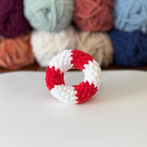 May include: A handmade, plush, donut-shaped toy in red and white. The toy is made of soft, textured yarn and has a circular shape with alternating red and white segments. The background features various balls of yarn in different colours.