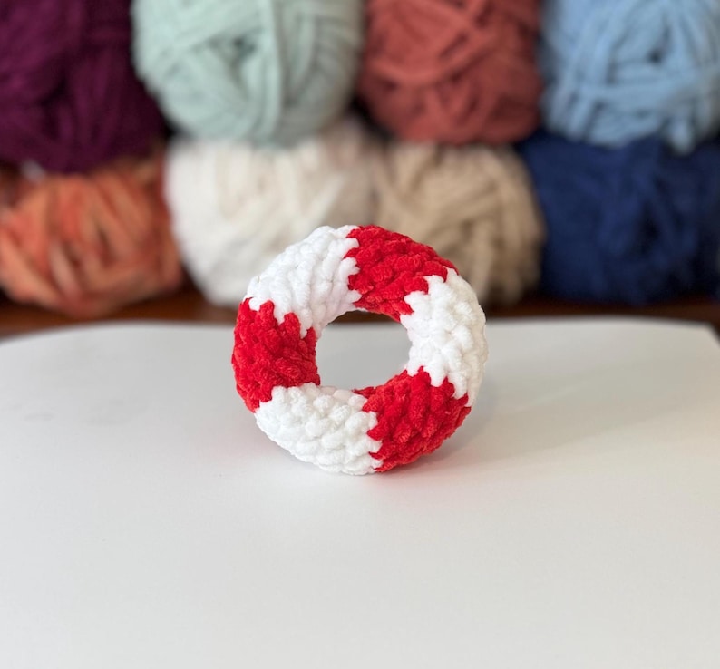 May include: A handmade, plush, donut-shaped toy in red and white. The toy is made of soft, textured yarn and has a circular shape with alternating red and white segments. The background features various balls of yarn in different colours.