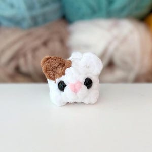 Puede incluir: Un peque&ntilde;o juguete de peluche tejido a mano en forma de cabeza de gato. La figura es principalmente blanca con una oreja marr&oacute;n, dos ojos redondos de pl&aacute;stico negro y una peque&ntilde;a nariz triangular de color rosa cosida en el centro.