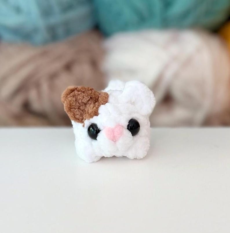Puede incluir: Un peque&ntilde;o juguete de peluche tejido a mano en forma de cabeza de gato. La figura es principalmente blanca con una oreja marr&oacute;n, dos ojos redondos de pl&aacute;stico negro y una peque&ntilde;a nariz triangular de color rosa cosida en el centro.