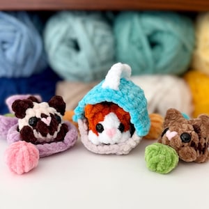Puede incluir: Tres peque&ntilde;os animales de peluche tejidos a ganchillo con hilo suave. Uno es una figura tipo pug marr&oacute;n oscuro y blanco sobre una base lavanda, otro es una criatura naranja y blanca dentro de una capucha azul, y otro es un gato marr&oacute;n con una bola verde.
