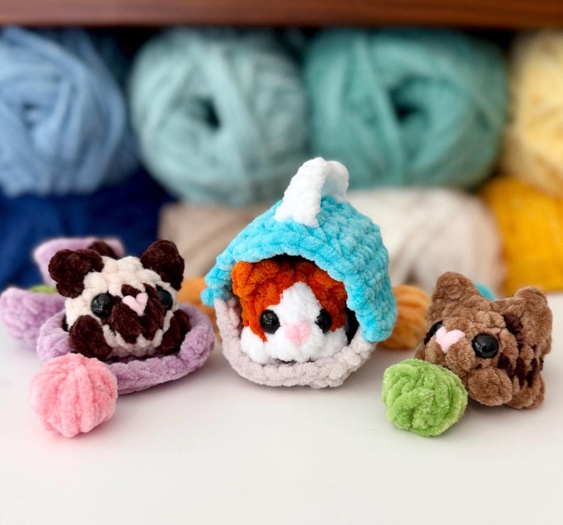 Puede incluir: Tres peque&ntilde;os animales de peluche tejidos a ganchillo con hilo suave. Uno es una figura tipo pug marr&oacute;n oscuro y blanco sobre una base lavanda, otro es una criatura naranja y blanca dentro de una capucha azul, y otro es un gato marr&oacute;n con una bola verde.