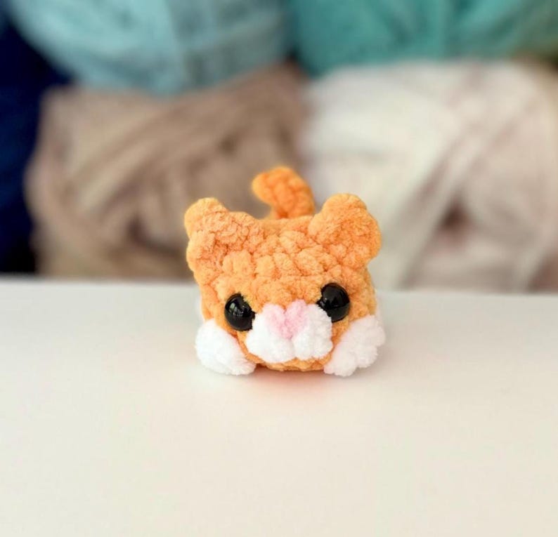 Puede incluir: Un peque&ntilde;o gato de peluche tejido a ganchillo en color naranja con una textura suave y esponjosa. El gato presenta dos ojos grandes y redondos de pl&aacute;stico negro, una peque&ntilde;a nariz triangular rosa y detalles de hilo blanco en el hocico y las patas.