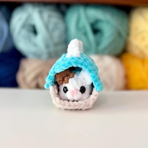 Puede incluir: Un peque&ntilde;o juguete de peluche tejido a mano que representa un animal blanco y marr&oacute;n con ojos negros y nariz rosa, colocado dentro de una cesta de color beige claro con una cubierta azul turquesa brillante hecha de hilo suave y texturizado.
