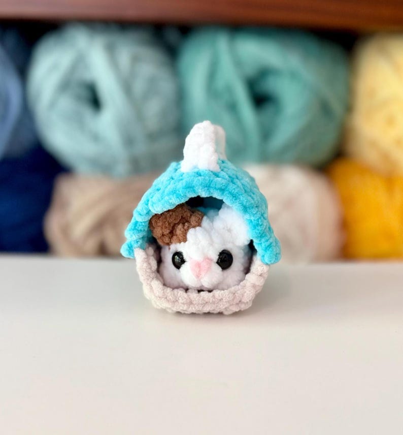 Puede incluir: Un peque&ntilde;o juguete de peluche tejido a mano que representa un animal blanco y marr&oacute;n con ojos negros y nariz rosa, colocado dentro de una cesta de color beige claro con una cubierta azul turquesa brillante hecha de hilo suave y texturizado.