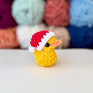 May include: A small, yellow crocheted duck wearing a red and white Santa hat. The duck has a small orange beak and black eyes. The background features a blurred array of colourful yarn skeins.