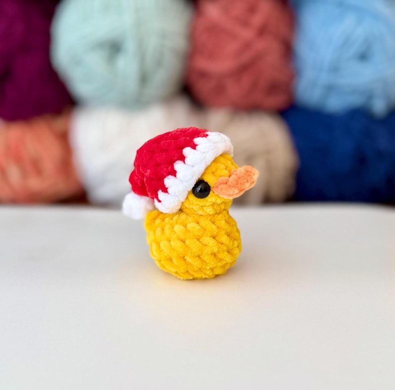 May include: A small, yellow crocheted duck wearing a red and white Santa hat. The duck has a small orange beak and black eyes. The background features a blurred array of colourful yarn skeins.